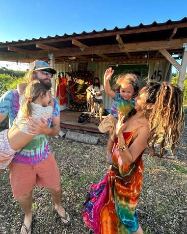 The image depicts a joyful family scene, with a man holding a child, a woman dancing, and a musician playing guitar on a porch, surrounded by a serene outdoor setting.