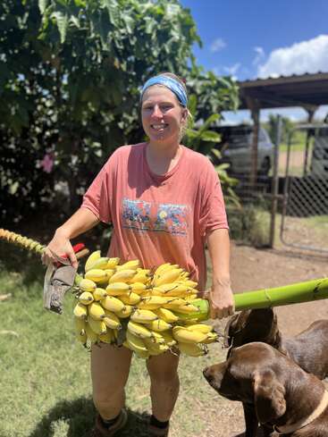 A woman stands in a yard, holding a large bunch of bananas and a machete, with a dog nearby, wearing a pink shirt and blue headband.