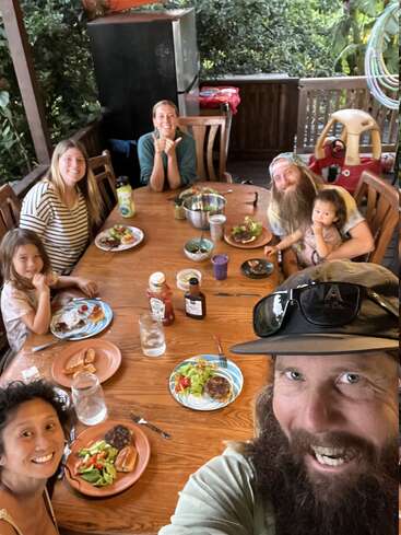 The image depicts a family of seven gathered around a wooden table, enjoying a meal together on a sunny day, surrounded by lush greenery.