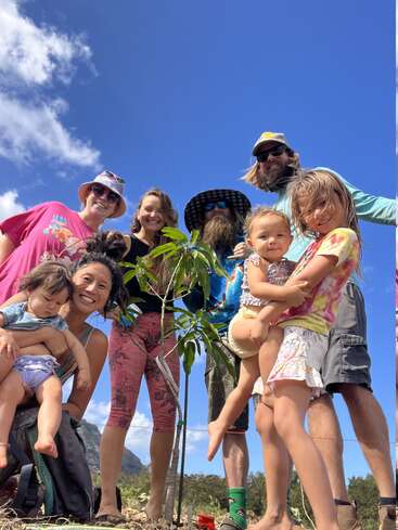 The image depicts a group of eight people, including children, posing for a photo with a small tree in front of a mountainous landscape on a sunny day.