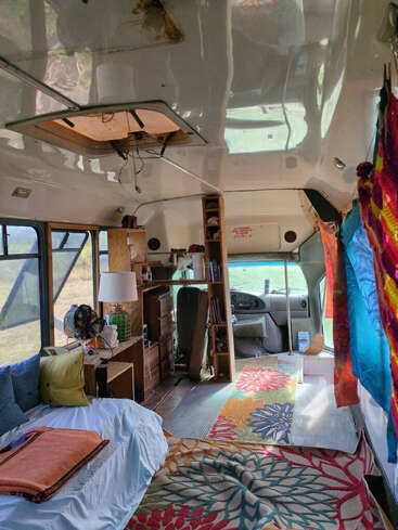 This image shows the cozy interior of a converted van or bus with a bed, colorful rugs, bookshelves, bright curtains, and natural light from windows.