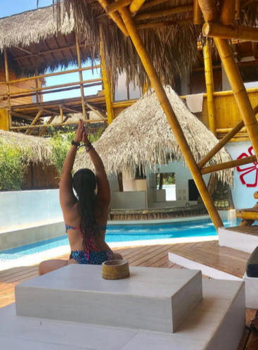 Una mujer en bikini se sienta junto a la piscina, con los brazos en alto en una postura de yoga, rodeada de bambú y estructuras con tejados de paja en un complejo turístico tropical.