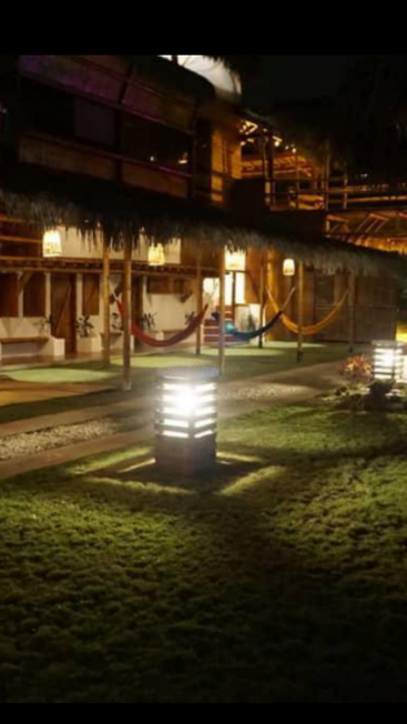 Una acogedora zona exterior cálidamente iluminada por la noche cuenta con hamacas, césped, farolillos modernos y un rústico edificio con tejado de paja, que crean un ambiente relajante y acogedor.