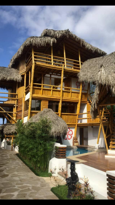 Esta imagen muestra un edificio tropical de bambú con tejados de paja, rodeado de vegetación, una pequeña piscina y un camino de piedra, bajo un cielo azul parcialmente nublado.