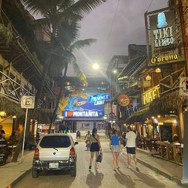 La imagen representa una vibrante escena callejera al atardecer, con palmeras, un coche blanco y peatones paseando por una calle de ladrillo bordeada de tiendas y restaurantes.