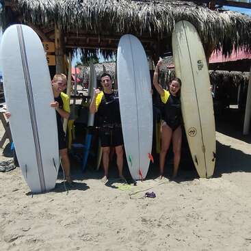Three people stand on a sunny, sandy beach, each holding a surfboard, smiling in front of a thatched-roof surf school hut under a blue sky.