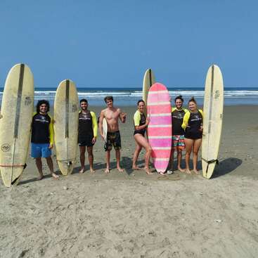 Seis personas sonríen en una playa de arena con una tabla de surf en la mano. Las olas del mar y el cielo azul claro de fondo sugieren un divertido día de surf.