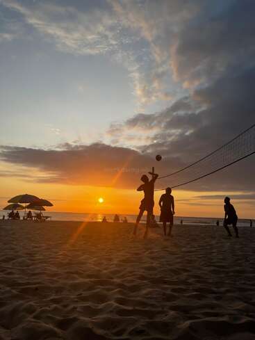 Silhouetted people play volleyball on a sandy beach at sunset, with vibrant orange sunlight, umbrellas, and clouds creating a peaceful yet lively evening atmosphere.
