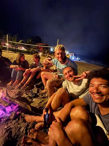 A group of friends sits barefoot on the sandy beach at night, gathered around a glowing bonfire, smiling and enjoying each other’s company under the night sky.