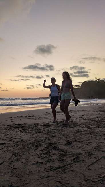 Two people walk barefoot along a sandy beach at sunset. One waves, the other carries sandals. The sky is pastel, dotted with clouds, creating a peaceful scene.