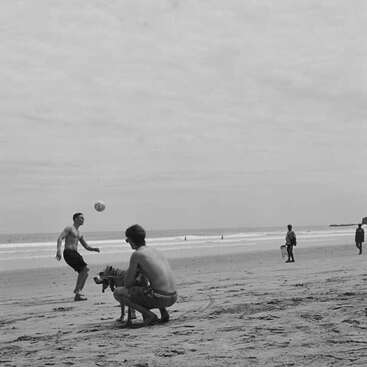Two shirtless men play with a ball and a dog on a sandy beach. Other people walk in the background. The scene is in black-and-white.