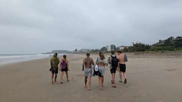 Sechs Leute spazieren an einem breiten, leeren Strand unter bewölktem Himmel entlang. Sie haben Freizeitkleidung an, tragen Taschen und genießen einen entspannten Tag am Meer.