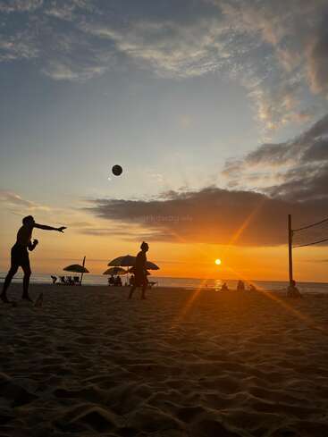 Eine wunderschöne Strandszene bei Sonnenuntergang, mit den Silhouetten von Leuten, die Volleyball spielen, Sonnenschirmen und ruhigen Wellen. Der leuchtende Himmel und die goldene Sonne sorgen für eine friedliche Atmosphäre.