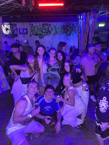 Eine Gruppe von Freunden macht zusammen auf einer coolen Party ein Foto. Die Stimmung ist super, mit blauen und violetten Lichtern, Drinks in der Hand und Leuten, die fröhlich lächeln.