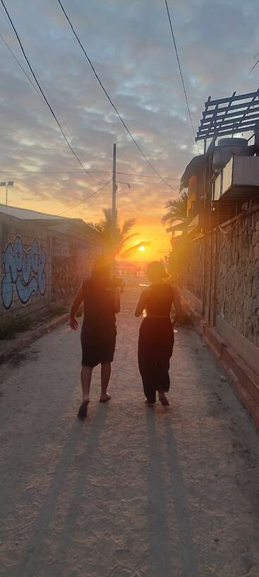 Zwei Leute laufen barfuß bei Sonnenuntergang eine sandige Gasse entlang und werfen lange Schatten, während warmes Licht leuchtet, umgeben von Graffiti-Wänden und Palmen.