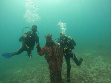 Zwei Taucher posieren unter Wasser neben einer coolen, mit Korallen bewachsenen Statue. Während sie lächeln, steigen Blasen über ihnen auf und zeigen eine abenteuerliche Unterwassererkundung im klaren blauen Wasser.
