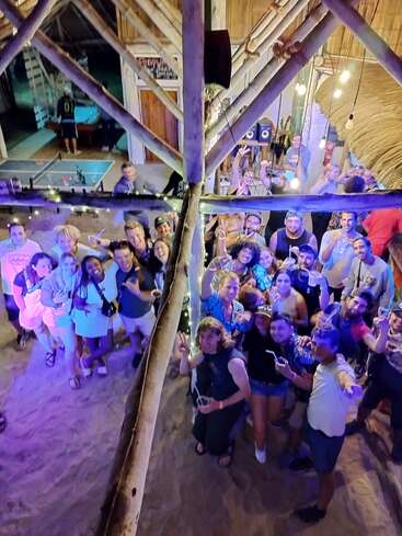 Eine fröhliche Gruppe von Leuten steht drinnen zusammen, lächelt und hat Drinks in der Hand, unter festlichen Lichtern und mit Holzbalken. Die Stimmung ist super, was auf eine Party oder Feier hindeutet.