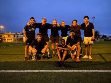 Acht Leute stehen in der Abenddämmerung auf einem Fußballfeld, tragen schwarze Shirts und Shorts, lächeln und sind total entspannt, was auf Kameradschaft und Teamwork nach einem Spiel hindeutet.