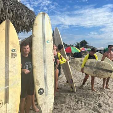 Un grupo de personas posa en una playa de arena con tablas de surf en las manos, algunas sonriendo, bajo sombrillas de colores, con palmeras y un cielo azul al fondo.