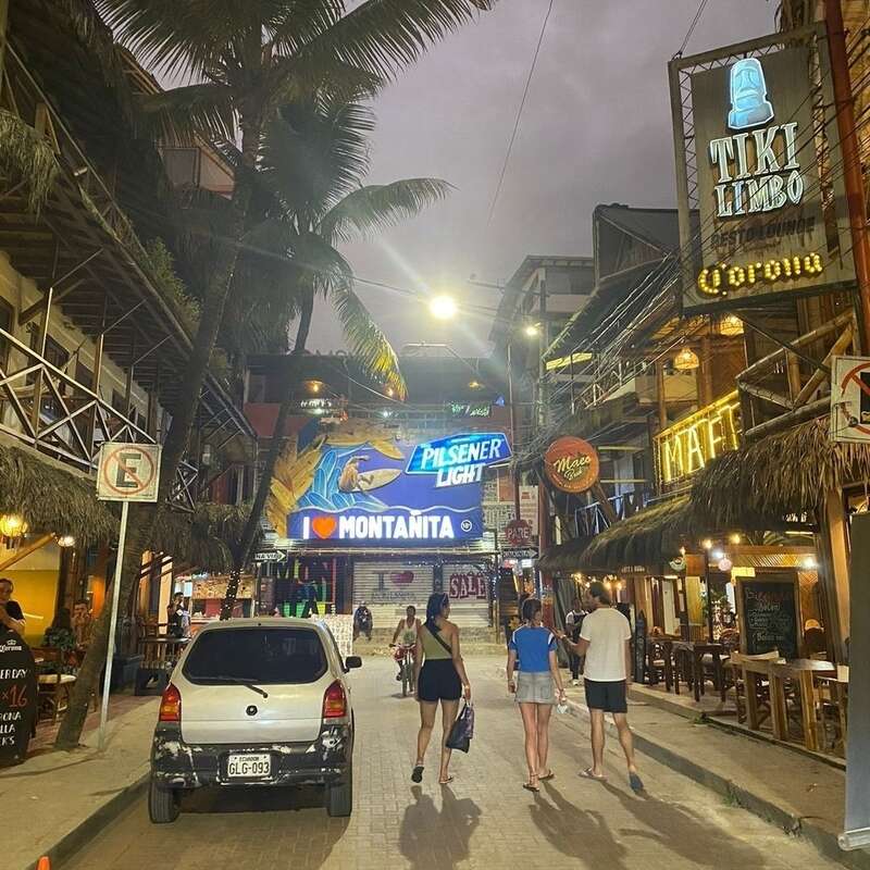 Das Bild zeigt eine lebhafte Straßenszene in der Abenddämmerung mit Palmen, einem weißen Auto und Fußgängern, die eine mit Ziegeln gepflasterte Straße mit Geschäften und Restaurants entlangschlendern.
