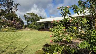 A charming house with a metal roof sits surrounded by lush green lawn, blooming flowers, vibrant shrubs, and mature trees under a partly cloudy sky.