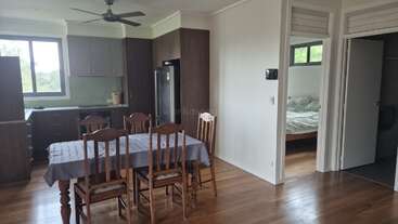 This image shows a kitchen and dining area with wooden floors, a table with six chairs, a ceiling fan, and adjacent bedroom and bathroom entrances.
