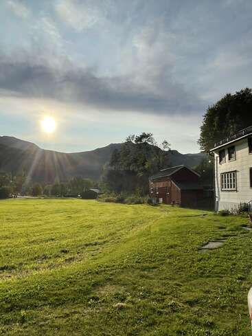 Um sol forte brilha sobre um campo verdejante, cercado por árvores, montanhas e edifícios rústicos. A cena é tranquila, campestre e banhada por uma luz dourada.