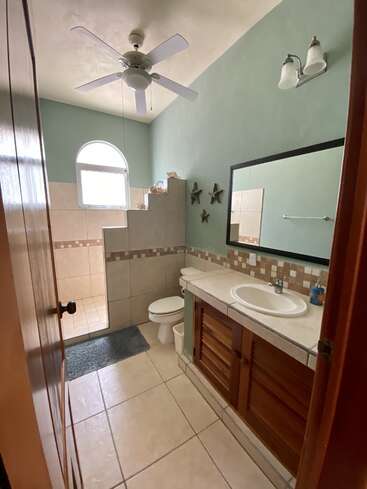 The image depicts a bathroom with a white sink, toilet, and shower, featuring light blue walls, a white ceiling, and beige tile flooring, with a window above the shower.