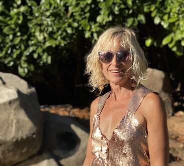 The image depicts a woman with blonde hair, wearing sunglasses and a gold sequined top, standing in front of a stone wall and greenery.