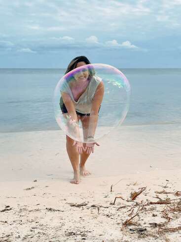 A woman stands on a beach, holding a large bubble in front of her, with the ocean and sky in the background, wearing a white shirt and black shorts.