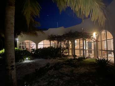 The image depicts a serene tropical house at night, featuring arched windows, a covered patio, and lush greenery, with a dark blue sky above.