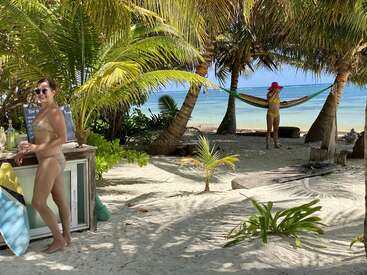 The image depicts two women on a tropical beach, with one standing at a bar and the other relaxing in a hammock between palm trees, enjoying the sun and sea.