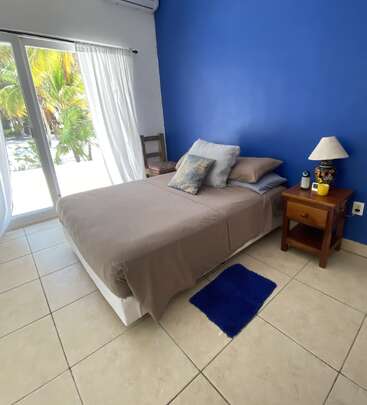 The image depicts a serene bedroom with a bed, nightstand, and sliding glass door leading to a patio with palm trees. The room is painted blue and white.