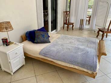 The image depicts a serene bedroom with a bed, nightstand, and lamp, featuring a white wall, tiled floor, and sliding glass doors leading to a patio area.