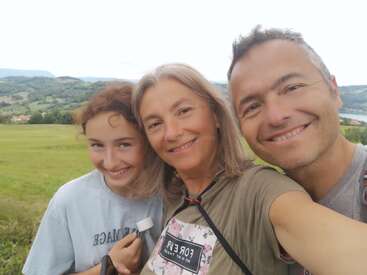A imagem mostra uma família de três pessoas posando para uma selfie em um campo pitoresco, com colinas e um corpo de água visíveis ao fundo.