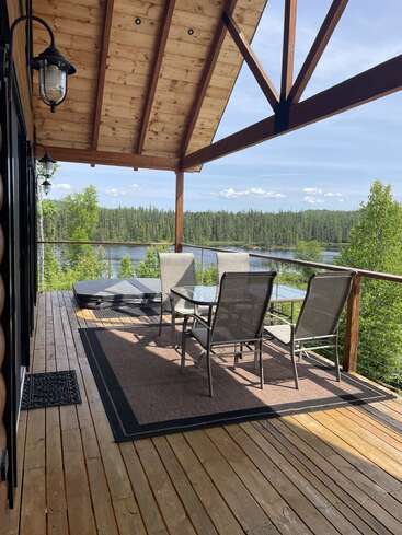 Una acogedora terraza de madera con techo abovedado, muebles de jardín y jacuzzi con vistas a un tranquilo lago y bosque bajo un cielo despejado y luminoso.