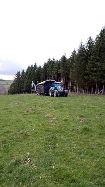 Un tracteur bleu tire une petite maison en bois et du matériel à travers un champ herbeux, près d'une forêt dense, sous un ciel clair et vaste, dans un cadre rural et paisible.