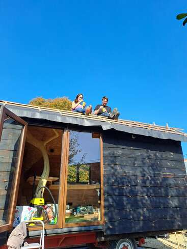 Deux personnes sont assises sur le toit d'une petite cabane en bois, dégustant des boissons sous un ciel bleu clair. L'intérieur de la cabane est confortable et accueillant.