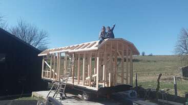 Deux personnes sont assises sur la charpente en bois d'une maison en construction sur une remorque, souriantes, en plein air, avec un ciel clair et des champs verts en arrière-plan.
