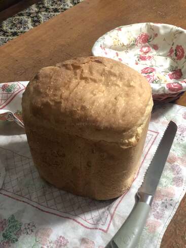 Um pão recém-assado está sobre um pano com estampa floral, ao lado de uma faca de pão e de uma tigela vazia com desenho de rosas, em cima de uma mesa de madeira.
