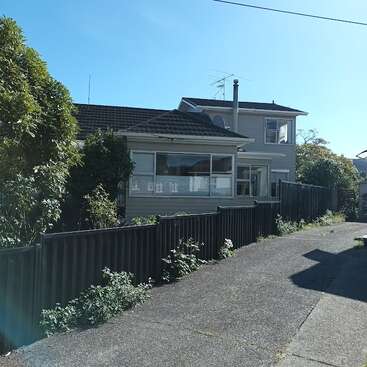Uma casa de dois andares com grandes janelas fica atrás de uma cerca de metal preta. Uma entrada de concreto leva você até lá, cercada por plantas e vegetação crescidas, sob um céu limpo.