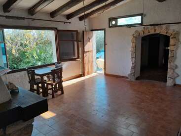 Habitación rústica con suelo de baldosas de terracota, vigas de madera en el techo, chimenea de piedra, pequeña mesa de comedor, puerta de piedra arqueada, ventanas abiertas y luz solar que entra por una puerta abierta.