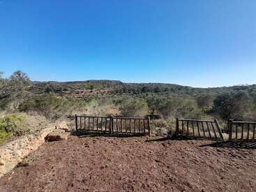 Un cielo azul despejado sobre un paisaje rural con colinas y árboles dispersos. En primer plano, una valla de madera rota, tierra seca y muros de piedra.
