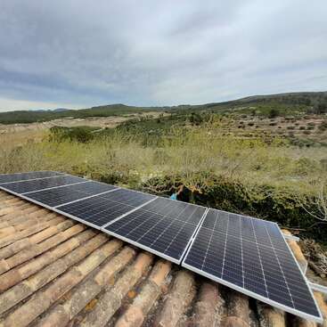 Esta imagen muestra unos paneles solares instalados en un tejado de tejas con vistas a un paisaje rural de colinas onduladas, vegetación verde y cielo nublado.