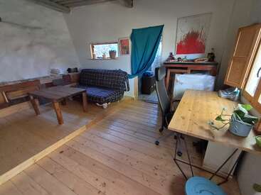 Esta acogedora habitación cuenta con suelos de madera, un sofá con funda a cuadros, una mesa rústica, un escritorio de madera con una planta, obras de arte, una cortina azul y luz natural.