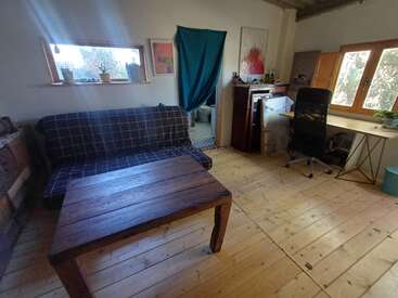 Una habitación acogedora e iluminada por el sol con un sofá a cuadros, una mesa de centro de madera, un escritorio con silla de oficina, plantas en macetas, suelos de madera clara y obras de arte decorativas.