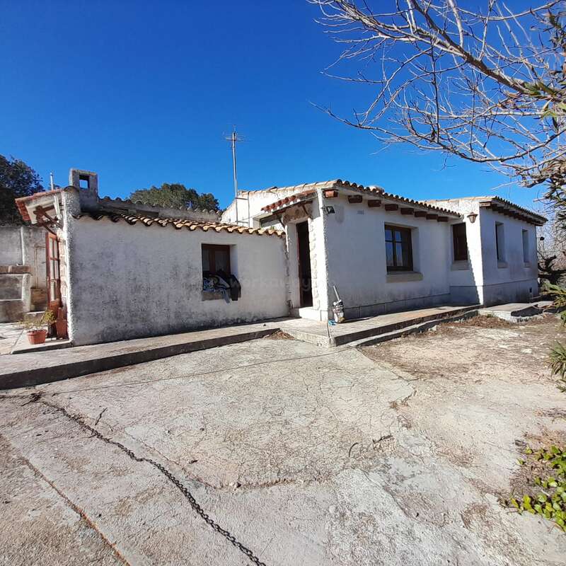 Das Bild zeigt ein kleines, weißes Haus mit Ziegeldach, das auf einer betonierten Auffahrt steht, umgeben von Bäumen und Büschen, vor einem klaren blauen Himmel.