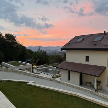 Una casa moderna con tejado rojo se alza en una ladera verde. El cielo al atardecer resplandece en tonos rosas y naranjas, con árboles y montañas lejanas a la vista.