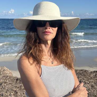 Eine Frau steht an einem sonnigen Strand und trägt einen weißen, breitkrempigen Hut, eine dunkle Sonnenbrille und ein gestreiftes Tank-Top. Wellen und blauer Himmel im Hintergrund.