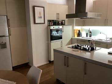 Cette cuisine moderne comprend des armoires de couleur crème, un îlot central avec une cuisinière à gaz, des appareils électroménagers en acier inoxydable, un four mural, des œuvres d'art, un éclairage épuré, des planchers en bois et un décor minimaliste.